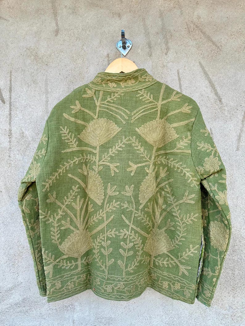 Op de afbeelding: Een groene jas met ingewikkelde bloemenborduurwerk in een lichtere groentint. De jas hangt aan een houten hanger tegen een neutraal gekleurde muur. De borduursels bedekken de achterkant van de jas.