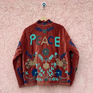 Pode incluir: Um casaco de veludo cor de ferrugem com a palavra PEACE bordada em letras grandes nas costas. O design inclui motivos florais, folhas e pássaros em tons de turquesa, azul escuro e bege, com gola alta e mangas compridas.