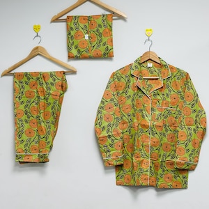 Op de afbeelding: Een groene pyjamaset met een oranje bloemenpatroon, bestaande uit een overhemd met knopen, een broek en een opgevouwen set. De pyjama's worden op houten kleerhangers tegen een witte achtergrond getoond. Het bloemenpatroon heeft oranje bloemen en groene bladeren.