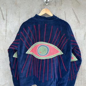 May include: A dark blue velvet jacket featuring a large embroidered eye design on the back. The eye has a green iris, a blue pupil, and a pink sclera, surrounded by radiating red embroidered lines that extend onto the sleeves of the garment.