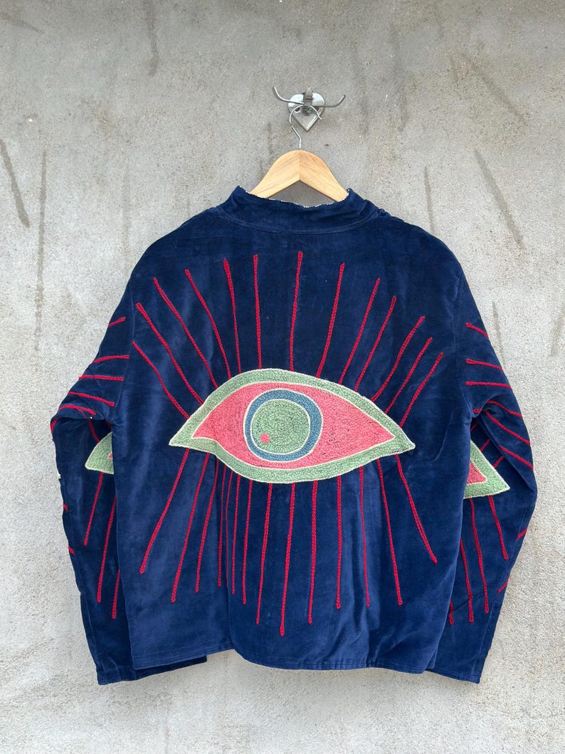 May include: A dark blue velvet jacket featuring a large embroidered eye design on the back. The eye has a green iris, a blue pupil, and a pink sclera, surrounded by radiating red embroidered lines that extend onto the sleeves of the garment.