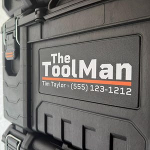 May include: Close-up of a black toolbox with the text "The ToolMan" in white letters and an orange line. The toolbox has a rectangular label with the name and phone number. The toolbox is made of a durable material.