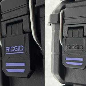 Puede incluir: Primer plano de un cierre de malet&iacute;n de herramientas negro con el nombre de la marca "RIDGID" en letras blancas y dos rayas horizontales moradas. El cierre est&aacute; hecho de un material duradero y texturizado y tiene una bisagra de metal plateado.
