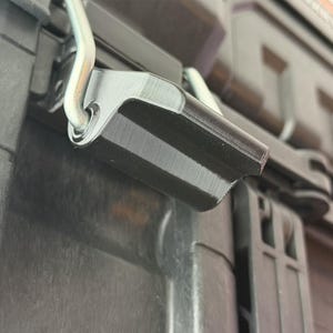 Puede incluir: Primer plano de un pestillo de pl&aacute;stico negro con una bisagra de metal plateado. El pestillo forma parte de un contenedor negro m&aacute;s grande, con crestas visibles y una superficie texturizada. La imagen est&aacute; bien iluminada.