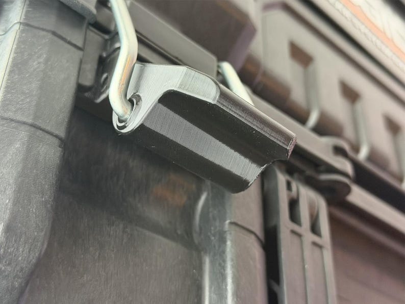 Puede incluir: Primer plano de un pestillo de pl&aacute;stico negro con una bisagra de metal plateado. El pestillo forma parte de un contenedor negro m&aacute;s grande, con crestas visibles y una superficie texturizada. La imagen est&aacute; bien iluminada.