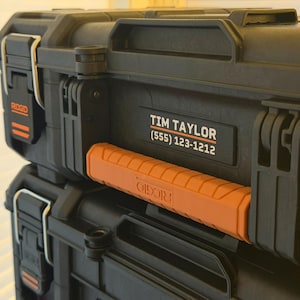 May include: Two black toolboxes stacked on top of each other. The top toolbox has a handle and the name "Tim Taylor" and a phone number. The handle is orange. The brand name "RIDGID" is visible on the side of the top toolbox.