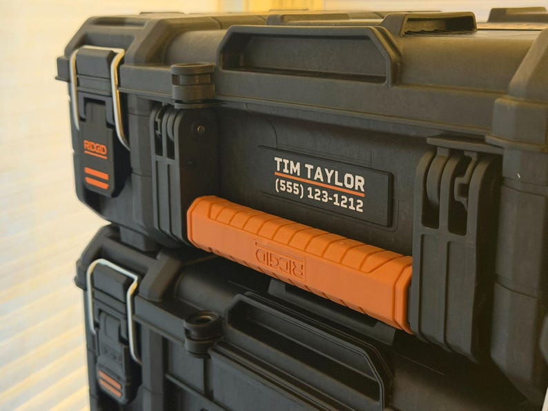 May include: Two black toolboxes stacked on top of each other. The top toolbox has a handle and the name "Tim Taylor" and a phone number. The handle is orange. The brand name "RIDGID" is visible on the side of the top toolbox.