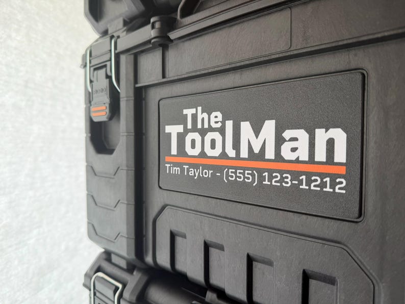 以下が含まれることがあります： 白い文字とオレンジ色の線で「The ToolMan」と書かれた黒いツールボックスのクローズアップ。 ツールボックスには、名前と電話番号が記載された長方形のラベルが付いています。 ツールボックスは耐久性のある素材で作られています。