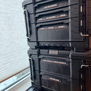 Puede incluir: Cajas de herramientas negras RIDGID Pro Gear System apiladas. Las etiquetas dicen: NUT ROUNDERS, LESS < ADJUSTERS > MORE, GUNS | DRUGS | MONEY, TIM TAYLOR (555) 123-1212, UGGA DUGGA & LIQUID WRENCH y THE BIG ONE.