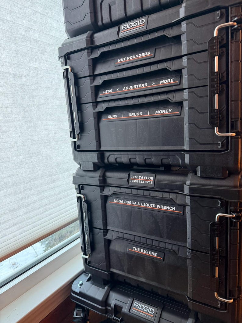Puede incluir: Cajas de herramientas negras RIDGID Pro Gear System apiladas. Las etiquetas dicen: NUT ROUNDERS, LESS < ADJUSTERS > MORE, GUNS | DRUGS | MONEY, TIM TAYLOR (555) 123-1212, UGGA DUGGA & LIQUID WRENCH y THE BIG ONE.