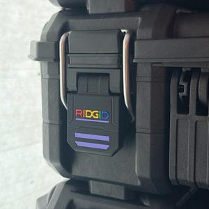 Puede incluir: Primer plano de una caja de herramientas negra con un cierre seguro. El cierre presenta el logotipo "RIDGID" en texto colorido. La caja tiene una superficie texturizada y un asa resistente.