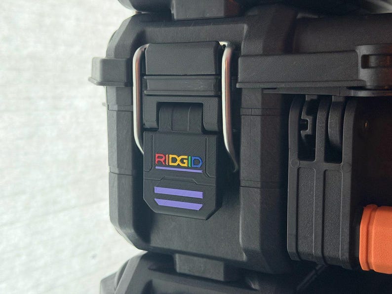 Puede incluir: Primer plano de una caja de herramientas negra con un cierre seguro. El cierre presenta el logotipo "RIDGID" en texto colorido. La caja tiene una superficie texturizada y un asa resistente.