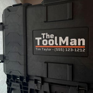 以下が含まれることがあります： 白とオレンジ色の「The Tool Man」の文字が入った黒いツールケース。ケースにはハンドルと電話番号が記載されたラベルが付いています。工具や機器の保管用に設計されています。
