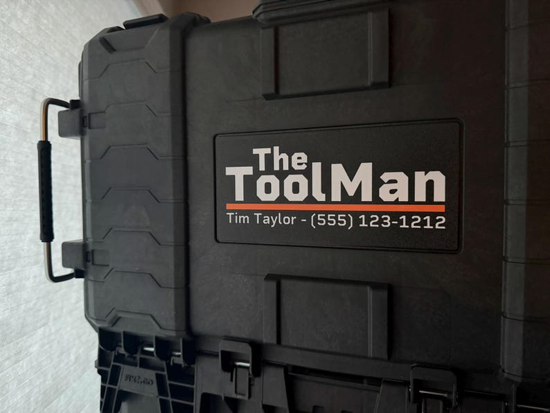 以下が含まれることがあります： 白とオレンジ色の「The Tool Man」の文字が入った黒いツールケース。ケースにはハンドルと電話番号が記載されたラベルが付いています。工具や機器の保管用に設計されています。