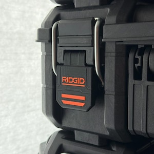 May include: Close-up of a black Ridgid tool case with a black latch and a silver hinge. The Ridgid logo is in orange. The case is made of a durable, textured material.