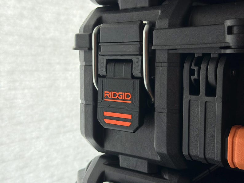 Puede incluir: Primer plano de una caja de herramientas negra Ridgid con un pestillo negro y una bisagra plateada. El logotipo de Ridgid es naranja. La caja est&aacute; hecha de un material duradero y texturizado.