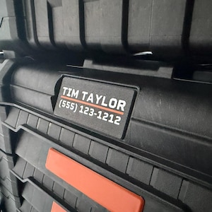 May include: Black storage container with a textured surface and orange accents. A label displays the name "TIM TAYLOR" and a phone number. The container is likely used for tools or equipment.