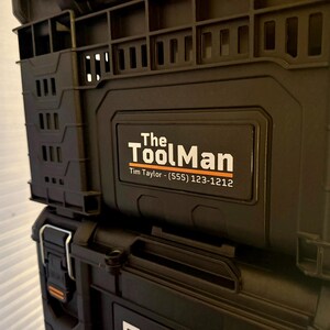Peut inclure: Conteneurs de rangement d'outils noirs empil&eacute;s. Le conteneur sup&eacute;rieur porte l'inscription "The ToolMan" en blanc et orange, avec un num&eacute;ro de t&eacute;l&eacute;phone en dessous. Les conteneurs ont une surface textur&eacute;e.