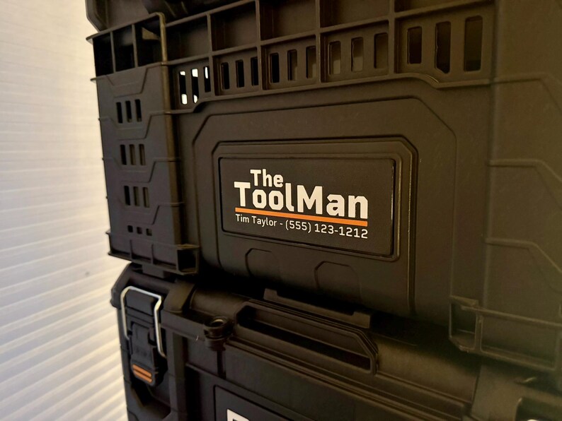 Peut inclure: Conteneurs de rangement d'outils noirs empil&eacute;s. Le conteneur sup&eacute;rieur porte l'inscription "The ToolMan" en blanc et orange, avec un num&eacute;ro de t&eacute;l&eacute;phone en dessous. Les conteneurs ont une surface textur&eacute;e.
