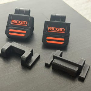 Puede incluir: Accesorios negros y naranjas para herramientas el&eacute;ctricas RIDGID. Dos piezas rectangulares negras con la palabra "RIDGID" en naranja y dos rayas naranjas. Tambi&eacute;n hay dos piezas negras en forma de L.