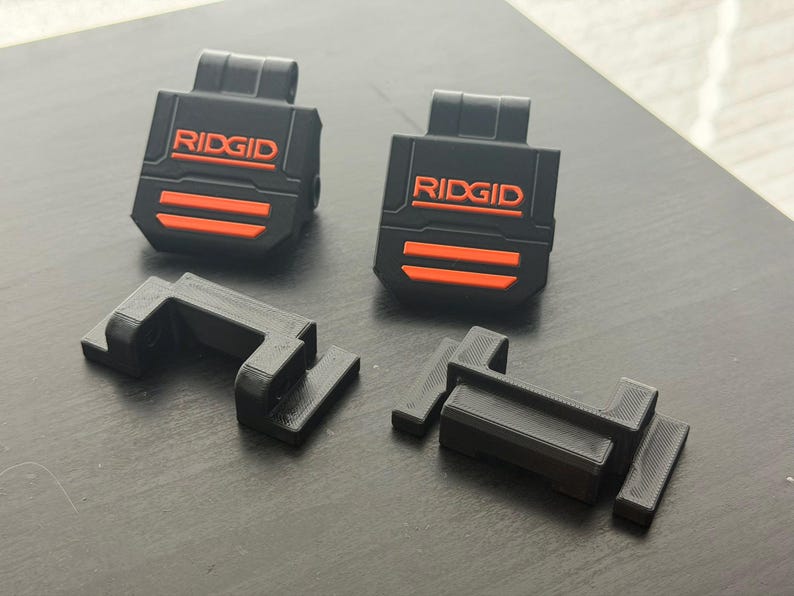 Puede incluir: Accesorios negros y naranjas para herramientas el&eacute;ctricas RIDGID. Dos piezas rectangulares negras con la palabra "RIDGID" en naranja y dos rayas naranjas. Tambi&eacute;n hay dos piezas negras en forma de L.