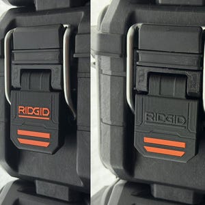 Puede incluir: Primer plano de un cierre de caja de herramientas negro con el nombre de la marca "RIDGID" en naranja. El cierre tiene dos rayas horizontales naranjas y un asa de metal plateado. La caja es negra.