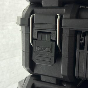Puede incluir: Primer plano de una caja de herramientas negra y resistente con un cierre met&aacute;lico plateado. El cierre se asegura con un cierre de pl&aacute;stico negro con el nombre de la marca "RIDGID" grabado. La caja est&aacute; dise&ntilde;ada para la durabilidad.