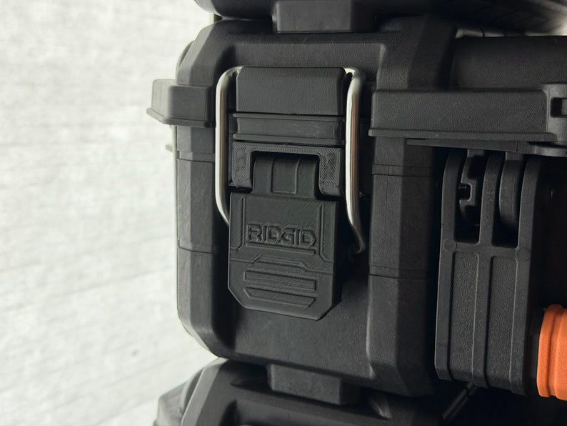 Puede incluir: Primer plano de una caja de herramientas negra y resistente con un cierre met&aacute;lico plateado. El cierre se asegura con un cierre de pl&aacute;stico negro con el nombre de la marca "RIDGID" grabado. La caja est&aacute; dise&ntilde;ada para la durabilidad.