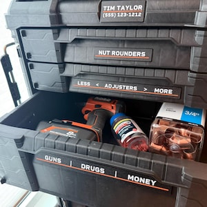 May include: Black toolbox with multiple drawers, labeled "NUT ROUNDERS", "LESS < ADJUSTERS > MORE", "GUNS", "DRUGS", and "MONEY". The open drawer contains a drill, a bottle of pills, and other items. The top drawer has the name "TIM TAYLOR" and a phone number.