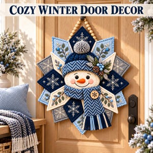 May include: A quilted star-shaped door hanging featuring a central snowman face with a carrot nose, wearing a blue chevron-patterned hat and scarf. The star is decorated with white snowflake embroidery, blue berries, and pine sprigs on a navy and light blue background.