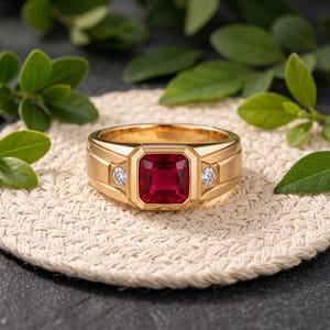 May include: A gold ring featuring a central square-cut deep red ruby flanked by two small round-cut clear diamonds. The band has a polished finish with a structured, geometric design surrounding the gemstones, creating a bold and symmetrical look.
