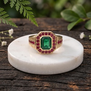 May include: A gold ring featuring a central emerald-cut green gemstone surrounded by a halo of small round red rubies. The band is crafted from polished yellow gold and extends into a wide, structured setting that frames the gemstone cluster.
