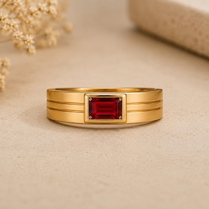 May include: A gold band ring featuring a rectangular, emerald-cut red gemstone set in the center. The ring has a smooth, matte finish with two horizontal grooved lines running along the sides of the band, framing the central red stone setting.