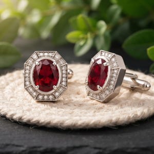 Peut inclure: Une paire de boutons de manchette argentés avec une pierre précieuse rouge foncé de taille ovale au centre, entourée d'un cadre géométrique octogonal incrusté de petites pierres claires. La monture est polie avec un fermoir à bascule.