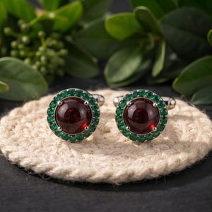 Peut inclure: Une paire de boutons de manchette ronds présentant une grosse pierre cabochon rouge foncé lisse au centre, entourée d'un halo de petites pierres vert vif serties dans une monture en métal argenté avec un mécanisme de fermeture à bascule.