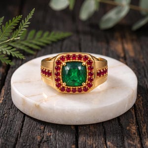 Pode incluir: Um anel de ouro com uma grande pedra preciosa verde de corte almofada no centro, rodeada por um halo de pequenas pedras vermelhas redondas. Pedras vermelhas pequenas adicionais estão cravadas na banda de ouro em ambos os lados do engaste quadrado.