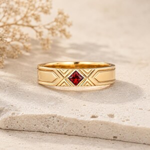 Könnte beinhalten: Ein goldener Ring mit einem zentralen, im Prinzessschliff geschliffenen roten Edelstein. Die Metalloberfläche weist ein geometrisches Muster aus eingravierten Linien und Winkeln auf, die den Stein umgeben und ein strukturiertes Design bilden.