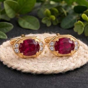 May include: A pair of gold cufflinks featuring large oval-cut deep red stones at the center, flanked by two small clear round stones on each side, set within a geometric gold frame with a polished finish and a standard T-bar toggle closure.