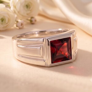 Pode incluir: Um anel de tom prateado com uma pedra preciosa de cor vermelho-escura e corte quadrado engastada no centro. A banda tem um acabamento liso e polido com detalhes laterais geométricos e estruturados que conduzem ao engaste quadrado.