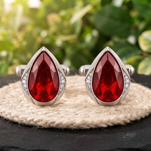 Peut inclure: Une paire de boutons de manchette argentés avec une grande pierre précieuse rouge facettée en forme de poire au centre, entourée d'une fine bordure de petites pierres claires étincelantes serties dans un cadre en métal poli avec fermeture.