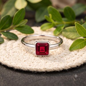 Könnte beinhalten: Ein silberfarbener Metallring mit einem quadratisch geschliffenen, tiefroten Edelstein in einer vierzackigen Fassung. Die Ringschiene ist glatt und poliert und zeigt ein klares Design, das die leuchtende Farbe des zentralen Steins hervorhebt.