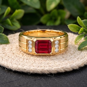 Könnte beinhalten: Ein goldener Ring mit einem großen rechteckigen roten Edelstein in der Mitte, flankiert von drei kleinen runden klaren Steinen auf jeder Seite. Die Ringschiene hat eine gebürstete Oberfläche mit horizontalen Rillen an den Seiten.