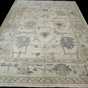 K&ouml;nnte beinhalten: Ein rechteckiger Teppich mit hellbeigem Hintergrund und einem sich wiederholenden Muster aus floralen und geometrischen Motiven in gedeckten Grau-, Salbeigr&uuml;n- und Braunt&ouml;nen, abgerundet durch eine Bord&uuml;re und Fransen.