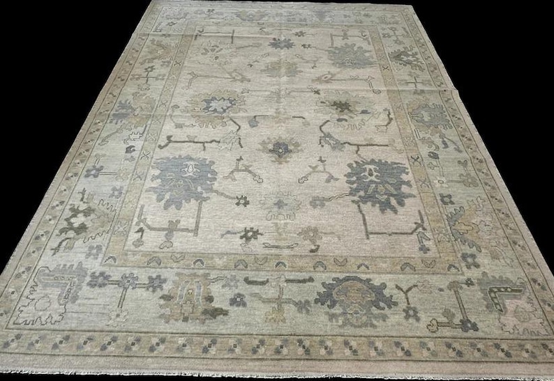 K&ouml;nnte beinhalten: Ein rechteckiger Teppich mit hellbeigem Hintergrund und einem sich wiederholenden Muster aus floralen und geometrischen Motiven in gedeckten Grau-, Salbeigr&uuml;n- und Braunt&ouml;nen, abgerundet durch eine Bord&uuml;re und Fransen.