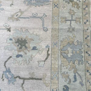 K&ouml;nnte beinhalten: Ein gewebter Teppich mit hellbeigem Hintergrund und gro&szlig;en, stilisierten Blumenmotiven in gedeckten T&ouml;nen von staubigem Blau, Salbeigr&uuml;n und Hellbraun. Die Bord&uuml;re zeigt ein sich wiederholendes geometrisches Muster mit blauen Akzenten.