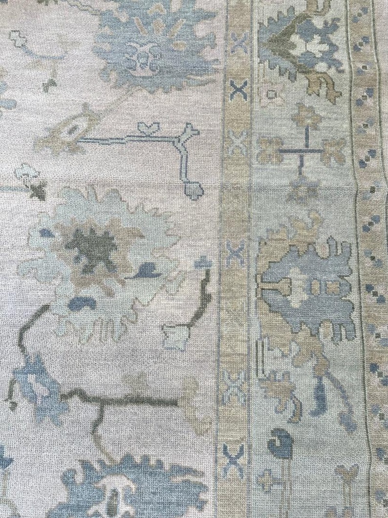 K&ouml;nnte beinhalten: Ein gewebter Teppich mit hellbeigem Hintergrund und gro&szlig;en, stilisierten Blumenmotiven in gedeckten T&ouml;nen von staubigem Blau, Salbeigr&uuml;n und Hellbraun. Die Bord&uuml;re zeigt ein sich wiederholendes geometrisches Muster mit blauen Akzenten.