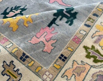 May include: A close-up of a hand-knotted rug with a light blue background and colorful geometric patterns. The rug features pink, yellow, green, and purple shapes, with a beige border. The text "HandKnottedRugiN" is visible.
