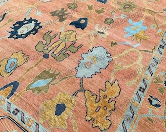 May include: Close-up of a peach-coloured rug with an intricate floral design. The rug features a repeating pattern of stylised flowers in shades of blue, yellow, and brown. The border has a geometric pattern in blue and light blue.
