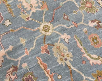 May include: Close-up of a hand-knotted rug with a floral pattern. The rug features a light blue background with a detailed design in shades of pink, green, and brown. The border has a cream and brown pattern.