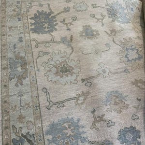 K&ouml;nnte beinhalten: Ein gro&szlig;er Teppich mit hellbeigem Hintergrund und einem wiederkehrenden Muster aus Blumen- und Rankenmotiven in gedeckten Schattierungen von Schieferblau, Taupe und sanftem Grau. Der Teppich hat eine breite Bord&uuml;re mit geometrischen Elementen.